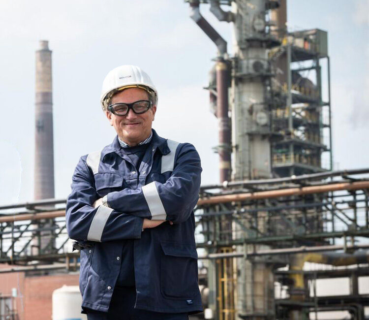 Ein Mann in blauer Arbeitskleidung und mit Helm steht mit verschränkten Armen vor einer chemischen Industrieanlage